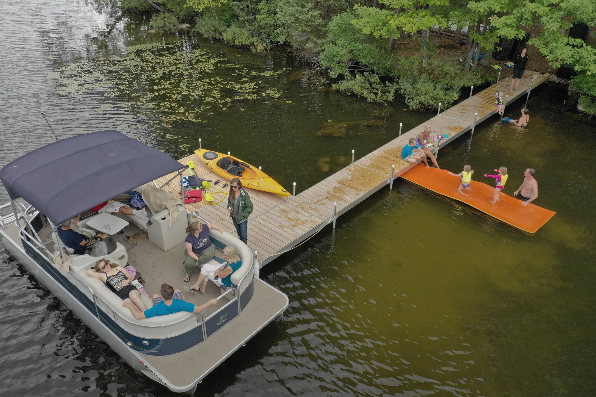 Pond scene with a pontoon boat, kayak, and dock with people around.